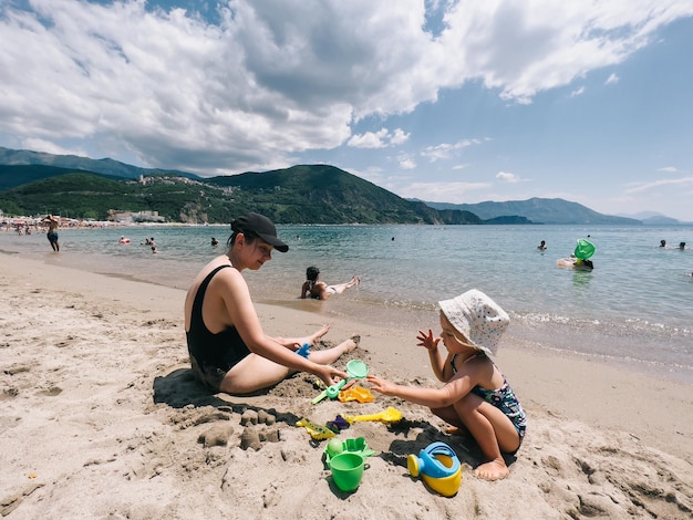 Мама с маленькой девочкой лепят фигурки из песка с пластиковыми формами на берегу моря
