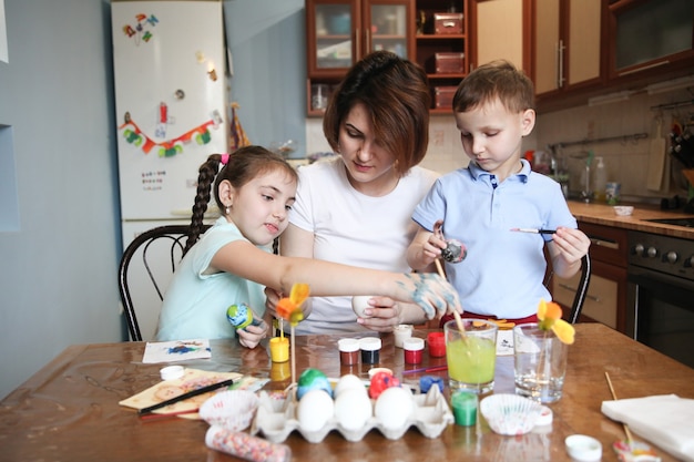 Мама с двумя детьми украшает пасхальные яйца, сидя за столом дома на кухне