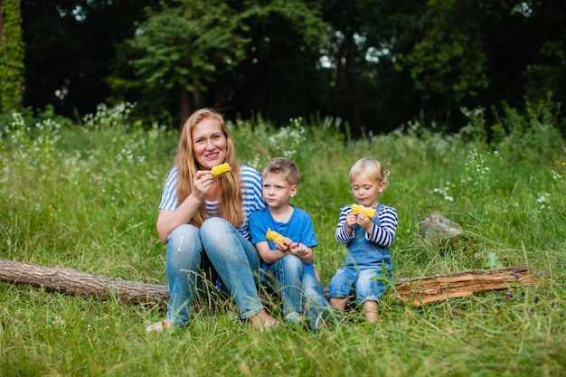 Mom with two children sitting in the woods on a fallen tree trunk and eating corn