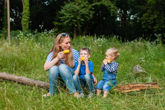 Mom with two children sitting in the woods on a fallen tree trunk and eating corn