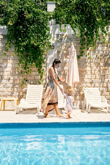 Mom with a watermelon in her hand leads a little girl to a sun lounger