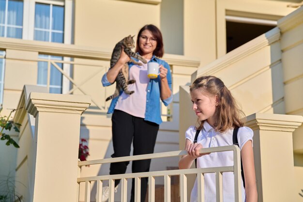 Mom withcat in arms on outdoor steps at home in morning with preteen daughter going to school