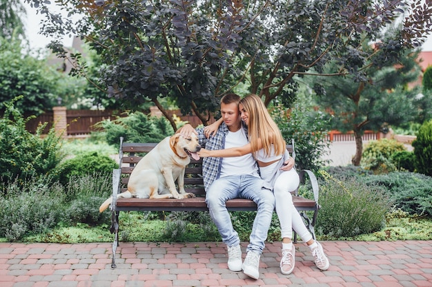 Il momento del riposo! bella coppia con il loro cane nel parco in una giornata di sole. giovane famiglia che accarezza il labrador e si siede sulla panchina.