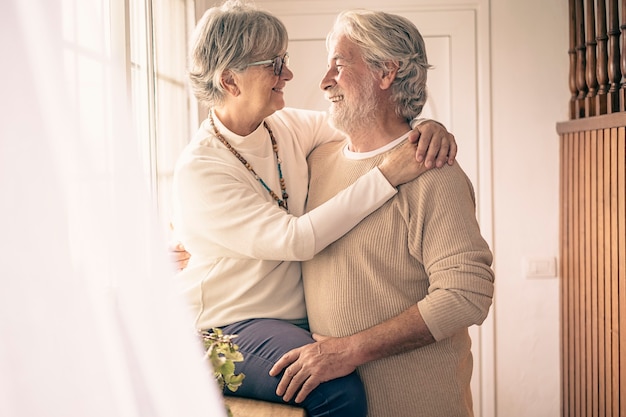 Un momento di tenerezza tra due persone anziane che si abbracciano con amore davanti alla finestra, guardandosi negli occhi. concetto di amore e felicità