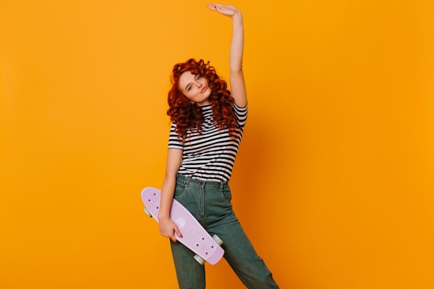 Momentopname van tienermeisje in goed humeur met paars skateboard in haar handen Roodharige dame in spijkerbroek poseren in oranje studio