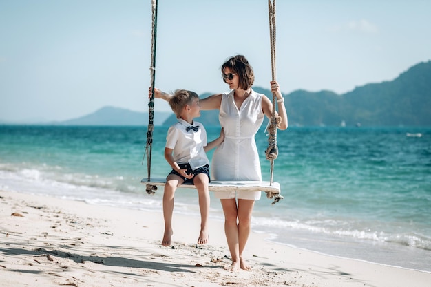 白いTシャツと蝶ネクタイを着た幼い息子と一緒に熱帯のビーチでスイングを楽しんでいるママ。プーケット。タイ。家族での休暇のコンセプト