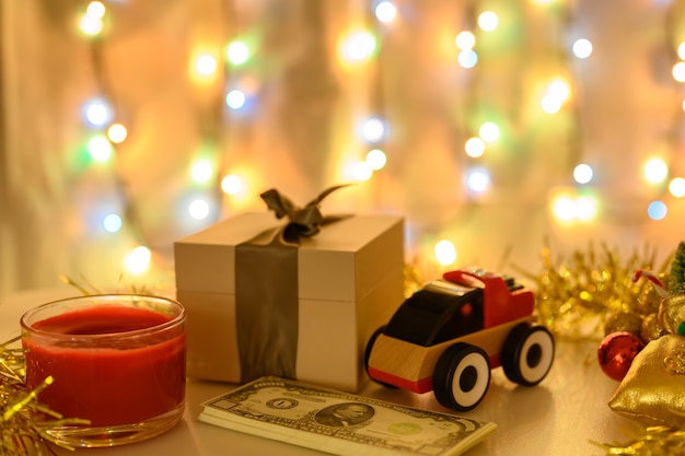 Soldi, una scatola con un regalo e una macchinina su uno sfondo natalizio con una ghirlanda luminosa dai colori caldi.
