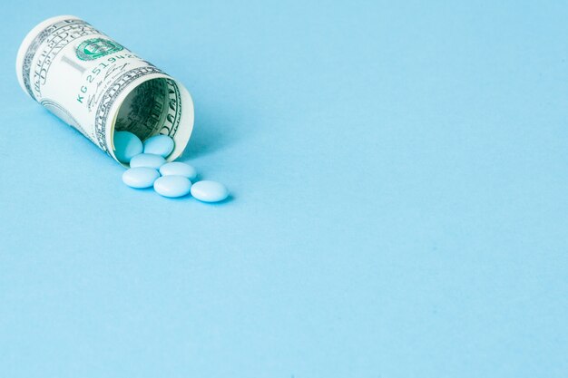 Money dollar rolled up with pills flowing out isolated on blue, high costs of expensive medication concept. Copy space