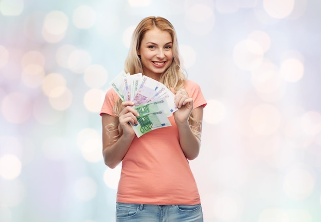 money, finances, investment, saving and people concept - happy young woman with euro cash money over blue holidays lights background