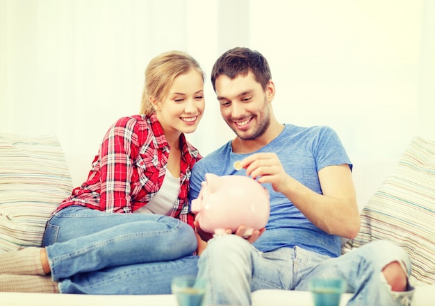 money, home, finance and relationships concept - smiling couple with piggybank sitting on sofa