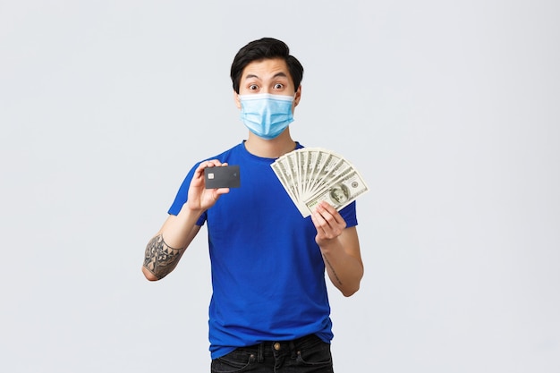 Money, lifestyle, insurance and investment concept. Excited good-looking young male student making deposit on banking account, showing credit card and cash dollars, stand in medical mask.