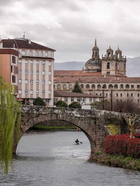 Monforte de Lemos Lugo 031322 A canoeist sailing surrounded by ducks on the waters of the river Cabe under a Roman stone bridge and the Nuestra Senora de la Antigua school in the background