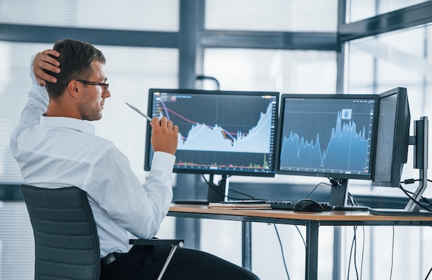 Monitoring information. Young businessman in formal clothes is in office with multiple screens. Conception of exchange and money.