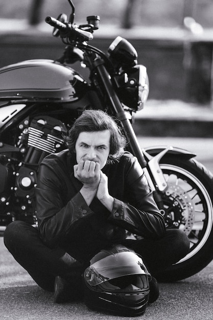 Монохромный портрет молодого человека в шлеме, сидящего возле мотоцикла байкерской концепции