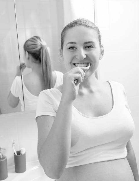 Monochrome portrait of young pregnant woman brushing teeth at bathroom