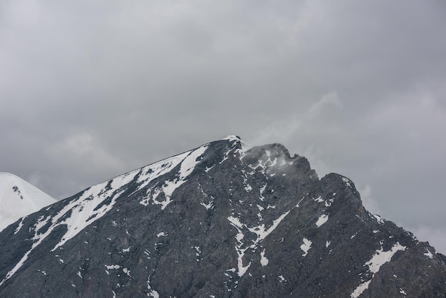 Monochrome scenery with high mountain peak with snow cornice in gray cloudy sky at overcast Dark atmospheric mountain landscape with large pinnacle in lead gray sky at rainy weather in grayscale