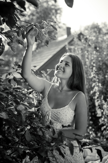 Monochroom schot van glimlachende vrouw die appel plukt van de top van de boom