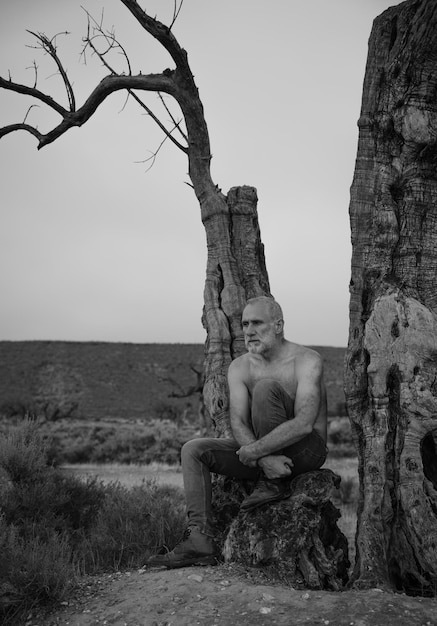 Foto monochroom van een shirtloze man die op een olijfboom tegen de lucht zit