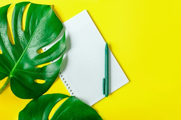 Monstera bladeren, een geruit notitieboekje en een pen op een trendy gele achtergrond. Ruimte kopiëren