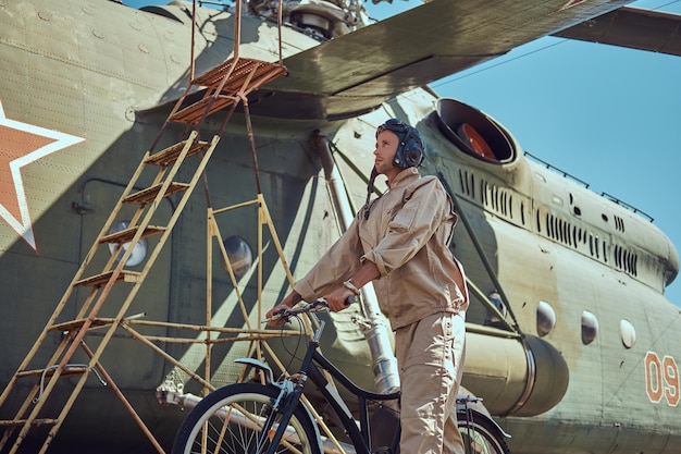 Monteur in uniform en vliegende helm wandelen met een fiets in de buurt van een grote militaire helikopter in een openluchtmuseum.