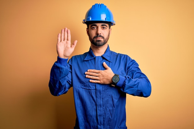 Monteur man met baard in blauw uniform en veiligheidshelm op gele achtergrond Vloekend met de hand op de borst en open handpalm die een eed van trouw aflegt