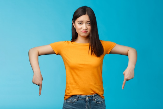 Moody sad asian brunette girl in yellow t-shirt smirking unsatisfied frowning bothered, complaining pointing down index fingers, showing bottom advertisement upset, jealous feel regret.