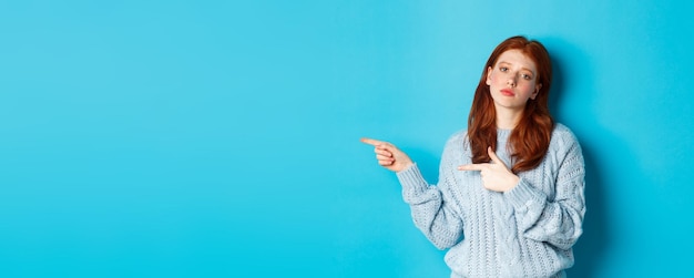 Moody teenage girl with red hair pointing fingers left at logo staring bothered and bored