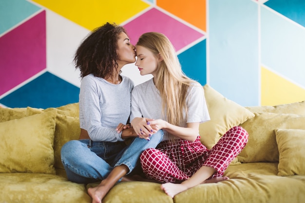 Mooi afrikaans amerikaans meisje met donker krullend haar kussen voorhoofd van mooi meisje met blond haar terwijl hand in hand elkaar met kleurrijke muur