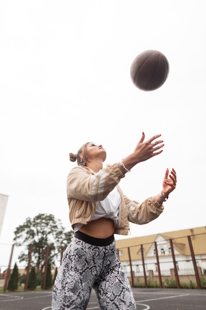 Mooi atletisch jong meisje in sportkleding met een windjack wit t-shirt en legging speelt basketbal en gooit de bal op straat