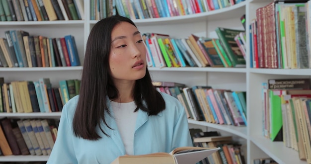 Mooi aziatisch meisje in blauw shirt loopt langs de planken met boeken in het onderwijsconcept van de bibliotheek college