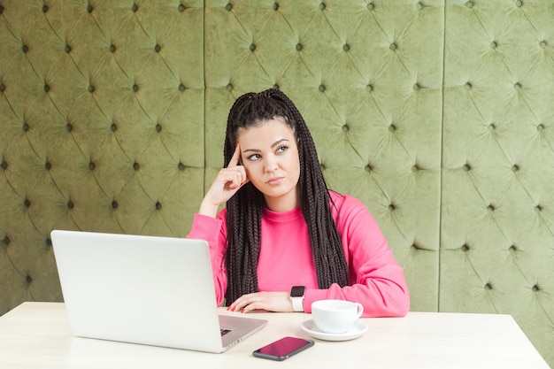 Mooi bedachtzaam jong meisje freelancer met zwarte dreadlocks kapsel in roze blouse zittend en werkend op laptop proberen nieuw idee te vinden en eigen strategie te plannen met vingers in de tempel