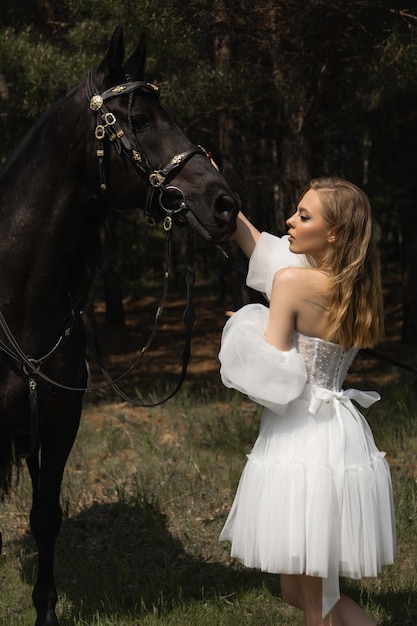 Foto mooi blank meisje in een trouwjurk die een paard in het gezicht streelt in het bos