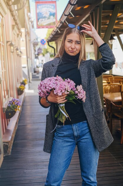 Foto mooi blank meisje met blond haar in een grijze jas en blauwe spijkerbroek met een boeket roze