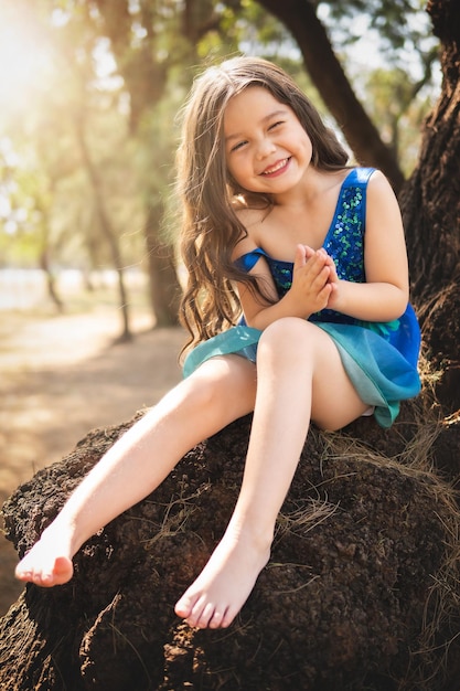 Foto mooi blij meisje spelen in het park in blauwe jurk zonder schoenen zitten in de boom