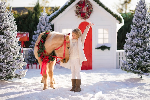 Mooi blond krullend kind en schattige pony met feestelijke krans bij het kleine houten huis en besneeuwde bomen.