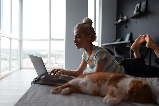 Foto mooi blond meisje dat thuis werkt en typt op laptop die op bed ligt in de buurt van rode kat