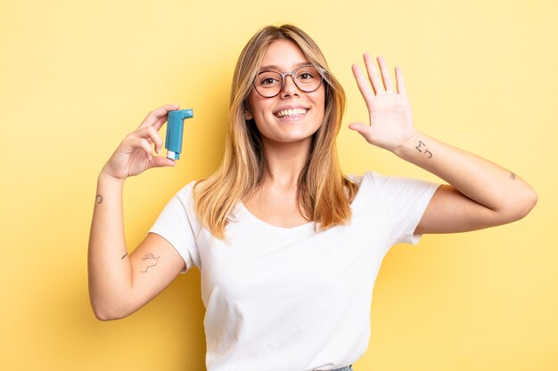 mooi blond meisje dat vrolijk lacht, met de hand zwaait, je verwelkomt en begroet. astma-inhalator concept