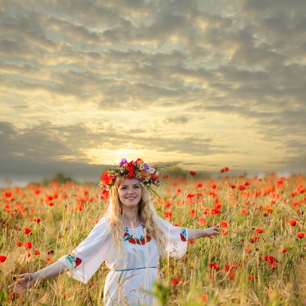 Mooi blond meisje in een jurk met een papaverpatroon en met een krans op zijn hoofd genietend in een veld met klaprozen