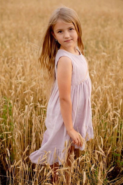 mooi blond meisje in een roze linnen jurk staat in een tarweveld en raakt haar aan met haar handen