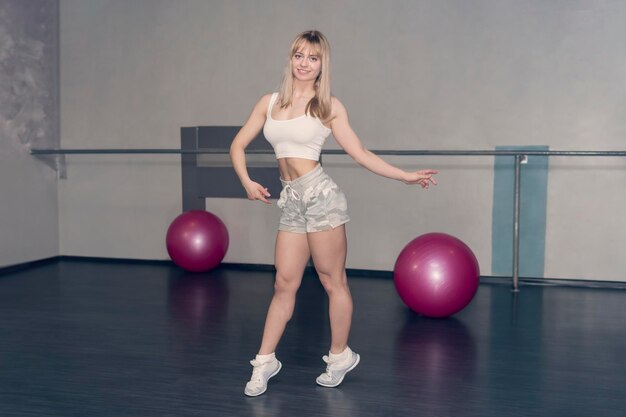 Mooi blond meisje in sportkleding doet oefeningen Glimlachend meisje in korte broek toont haar opgepompte spieren Schattig schattig meisje in de sportschool met roze ballen