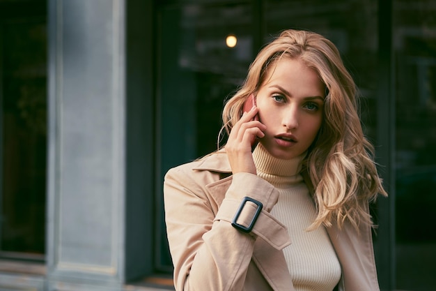 Mooi blond meisje in trenchcoat praten op mobiele telefoon sensueel in de camera kijken buiten