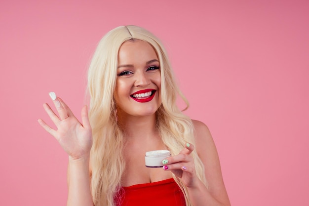 Mooi blond meisje met een heldere huid houdt de handen in de pot met crème in studio roze achtergrond.