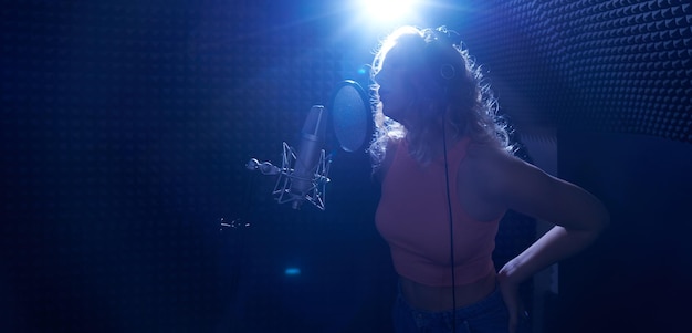 Mooi blond meisje zingt emotioneel lied in opnamestudio met professionele microfoon en koptelefoon creëert een nieuw trackalbum vocale artiest silhouet in blauw licht