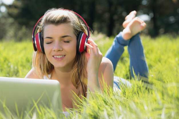 Mooi blonde die op gras liggen die laptop met behulp van die aan muziek op een zonnige dag in het platteland luisteren