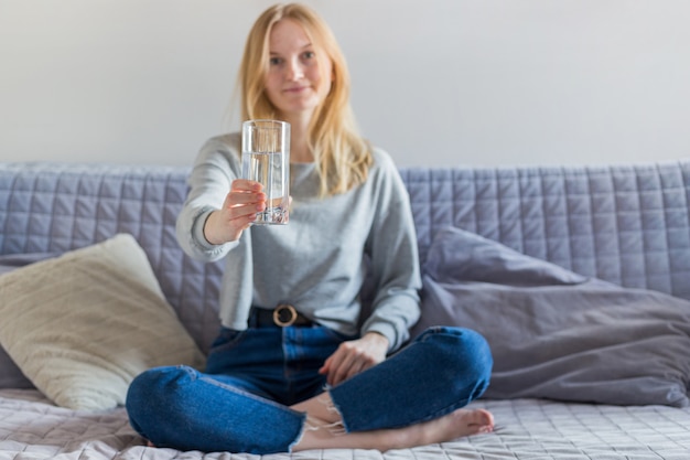 Mooi blondevrouw drinkwater en thuis het zitten op de bank in de woonkamer. Mooi jong meisje met glas water, goedemorgenconcept