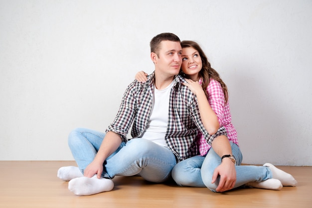 Mooi blootvoets jong paar in het toevallige jeans zitten die tegen een witte muur op de houten vloer in de woonkamer leunen die bij de camera glimlachen