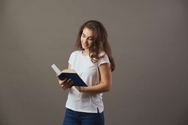 Mooi bruinharig meisje gekleed in een wit t-shirt en spijkerbroek leest een boek op een grijze achtergrond.