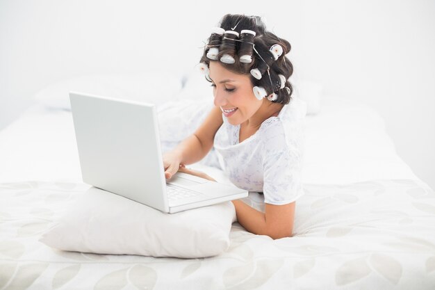 Mooi brunette in haarrollen die op haar bed liggen die haar laptop met behulp van