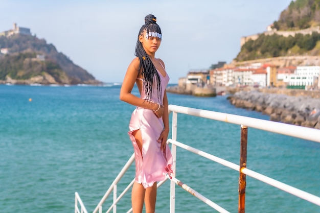 Mooi Dominicaanse etnische meisje met vlechten met een mooie roze jurk. Mode genieten van de zomer op een houten loopbrug aan zee