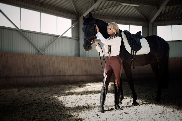 Mooi elegant jong blondemeisje die zich dichtbij haar paard bevinden die de eenvormige concurrentie kleden zich